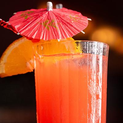 Close up of a tall glass filled with orange cocktail, and garnished with orange slice and umbrella.
