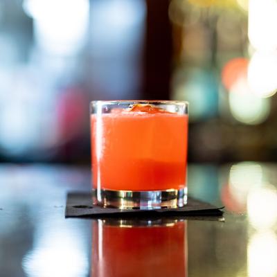 Golden Hour, Bright orange-red cocktail in rocks glass on blurred bar background.