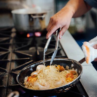Kitchen staff member preparing a dish in a frying pan with kitchen tongs