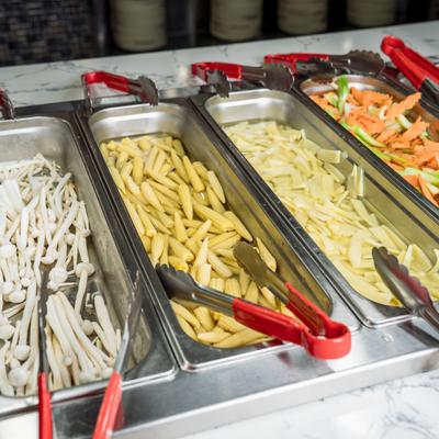 A buffet line features metal trays filled with various fresh, sliced vegetables.