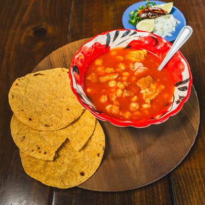 Bowl of Pozole soup served on a wooden board with tortillas on the side.