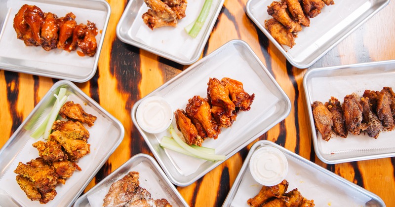 Multiple trays of assorted chicken wings with dipping sauces and celery on a wooden table