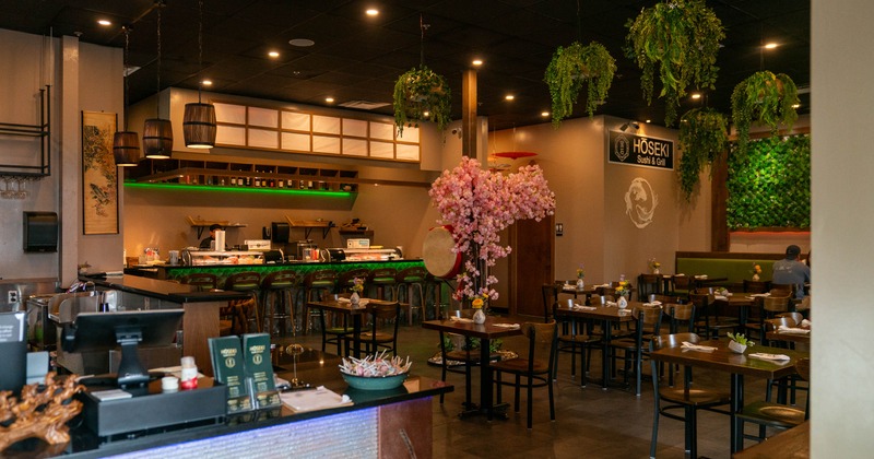 A warmly lit interior - a sushi bar, dining tables, hanging plants, pink floral decorations