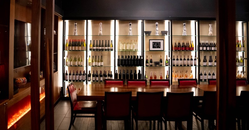 Seating area with long tables and chairs, glass wall shelves with wine  bottles