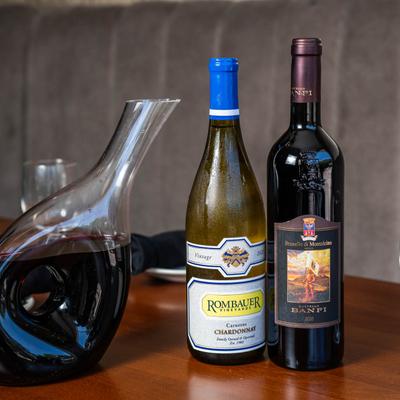 A glass decanter filled with red wine sits beside two wine bottles and a glass.