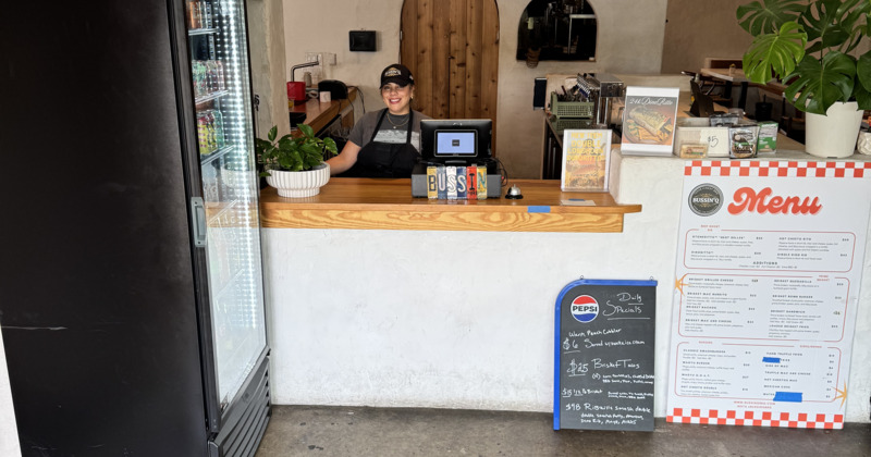 A smiling employee stands behind a wooden counter with a cash register