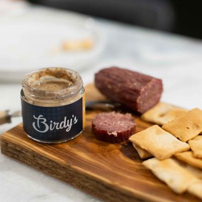 A tiny mustard jar with cured sausage and crackers on a wooden board.