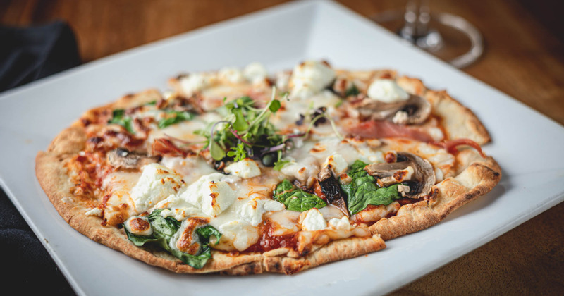 Pizza on a white plate features melted cheese, spinach, mushrooms, goat cheese, and microgreens