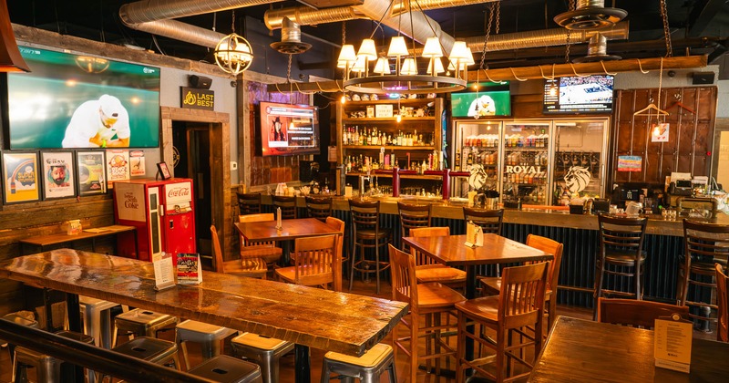 Warmly lit bar interior with wooden tables, metal stools, and multiple wall-mounted televisions