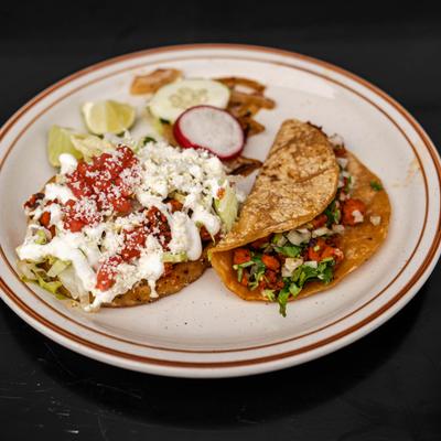 Sope topped with meat, lettuce, queso fresco, and crema, served alongside a taco.