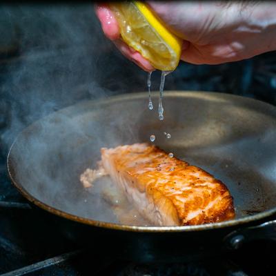 A hand squeezing a lemon above a sizzling salmon fillet in a pan.