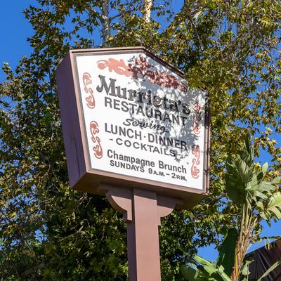 A business sign with the restaurant name and logo by a tree.