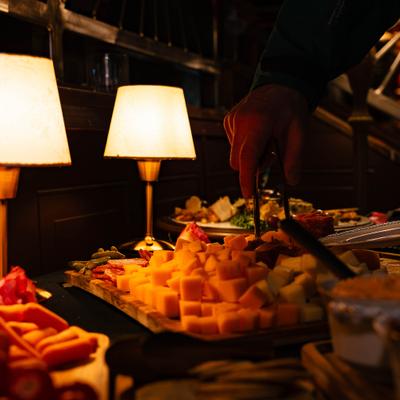 A warm-lit buffet scene shows a hand using tongs to pick cheese cubes from a platter.