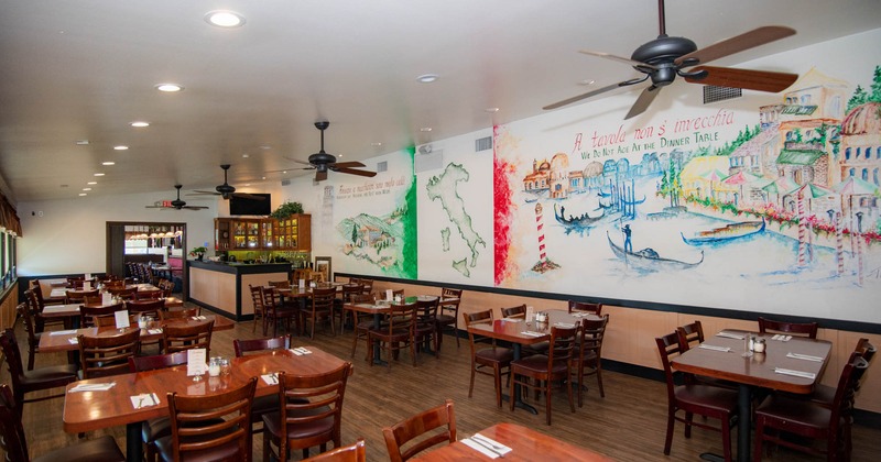 Interior of a restaurant with wooden tables, chairs, and colorful Italian-themed murals on the walls