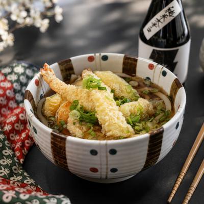 A steaming bowl of tempura udon sits next to sake and chopsticks.