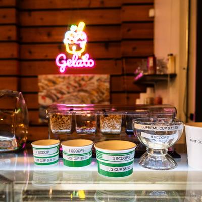 Pricing and menu options for a gelato counter, featuring a brightly lit neon sign.