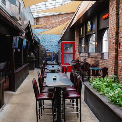 Cozy brick-walled patio with high tables and red chairs, adorned by greenery and TV screens.