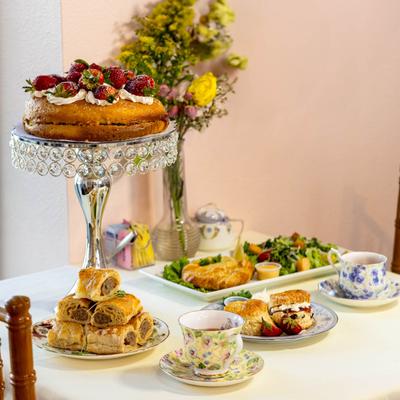 Strawberry-topped cake on a stand surrounded by sausage rolls, teacups, sandwiches, and flowers.