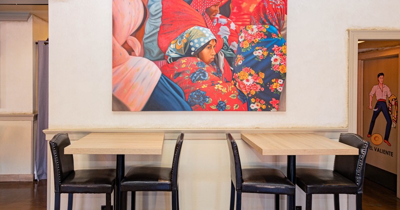 An interior with a colorful painting, two tables with black chairs in front