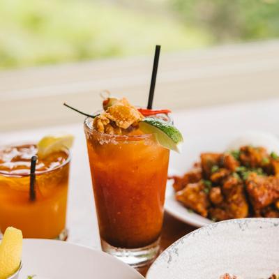Spicy Bloody Mary with savory garnish served alongside an orange cocktail and appetizers.
