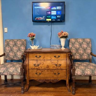 Vintage wooden dresser between two upholstered chairs beneath a wall-mounted TV.