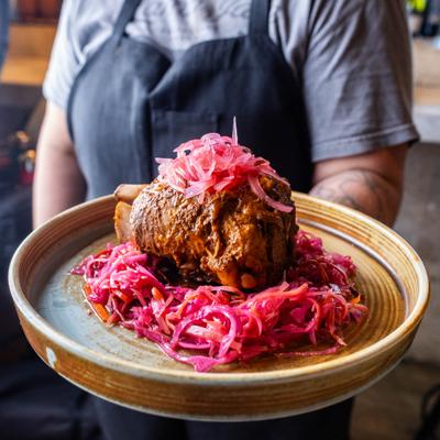 Server holding a plate with braised pork shank over spicy red slaw.