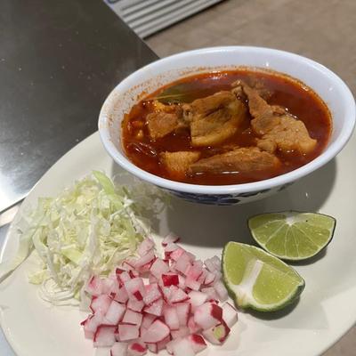 Bowl of Menudo soup served with onions, lettuce and lime wedges.