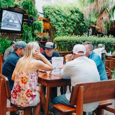 A group of guests sitting at a wooden table in an outdoor setting, surrounded by lush greenery.