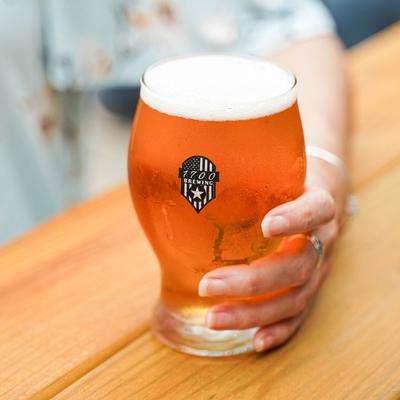 A hand holds a clear glass of amber beer with 1700 Brewing trademark.
