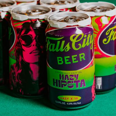 Vibrant cans of Falls City Beer on a green surface; colorful designs with stylized faces / text.