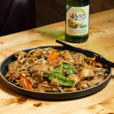 Drunken noodles with vegetables on a dark plate, accompanied with a soju bottle.