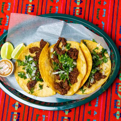 Carne Asada Tacos topped with onion and cilantro, served with lime wedges and a dip.