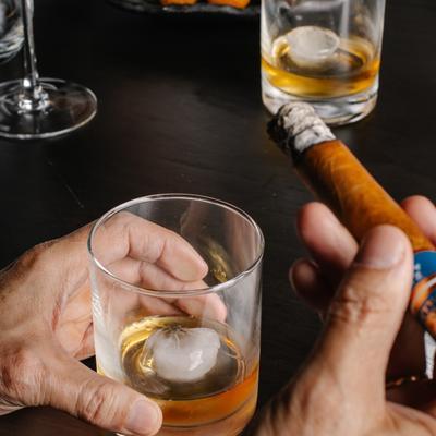 Guest holding an Old Fashioned and a cigar at a black table, with another drink behind.