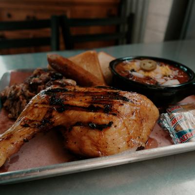 Barbecue platter with chicken leg, pulled pork, sausage, bread and small pot of chili.