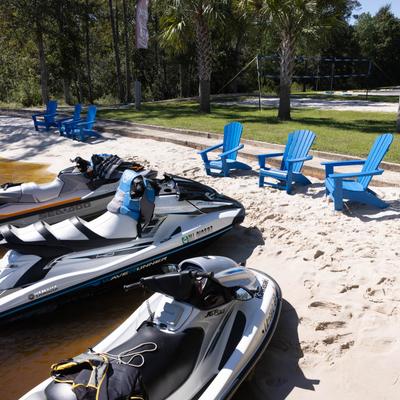Jet skis on the sand near blue chairs with trees in the background.
