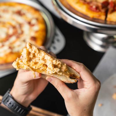 Hands holding a sandwich filled with creamy cheese topping, with pizzas in the background.