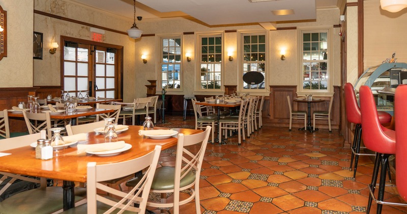 A brightly lit restaurant interior with terracotta tile flooring, wooden tables, and red bar stools