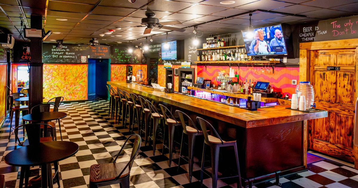 Bar interior with a long wooden counter, high stools, checkered floor, TVs, and vibrant decor