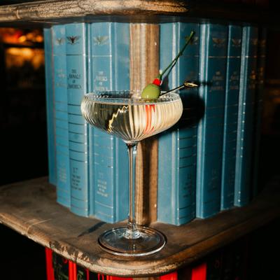 A cocktail glass with a martini and an olive garnish rests on a shelf of blue books.
