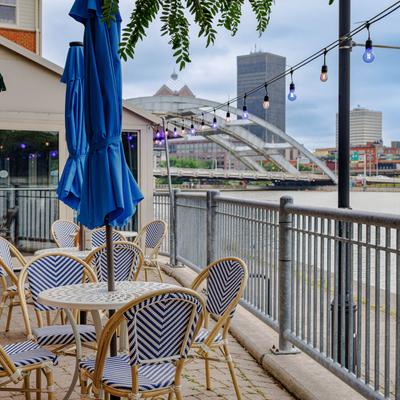 Riverside outdoor patio with blue umbrellas, string lights, and city bridge view.