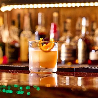 Amber cocktail on ice in rocks glass with orange and cherry garnish.