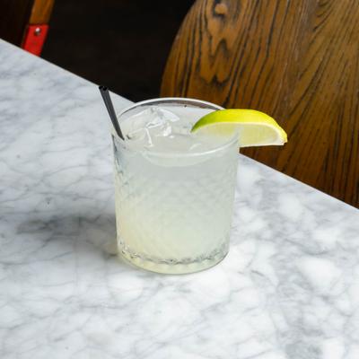 light yellow drink with a lime wedge on marble counter with wood chair in background.
