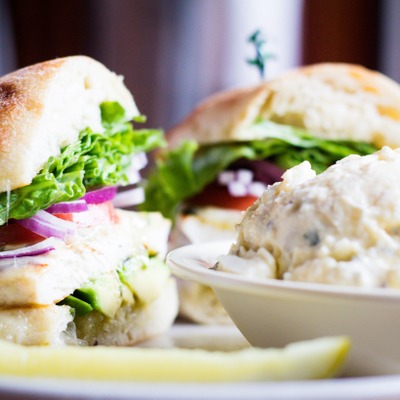 Chicken sandwich on a cibatta roll with lettuce, tomato and avocado inside. Plated next to a serving of potato salad