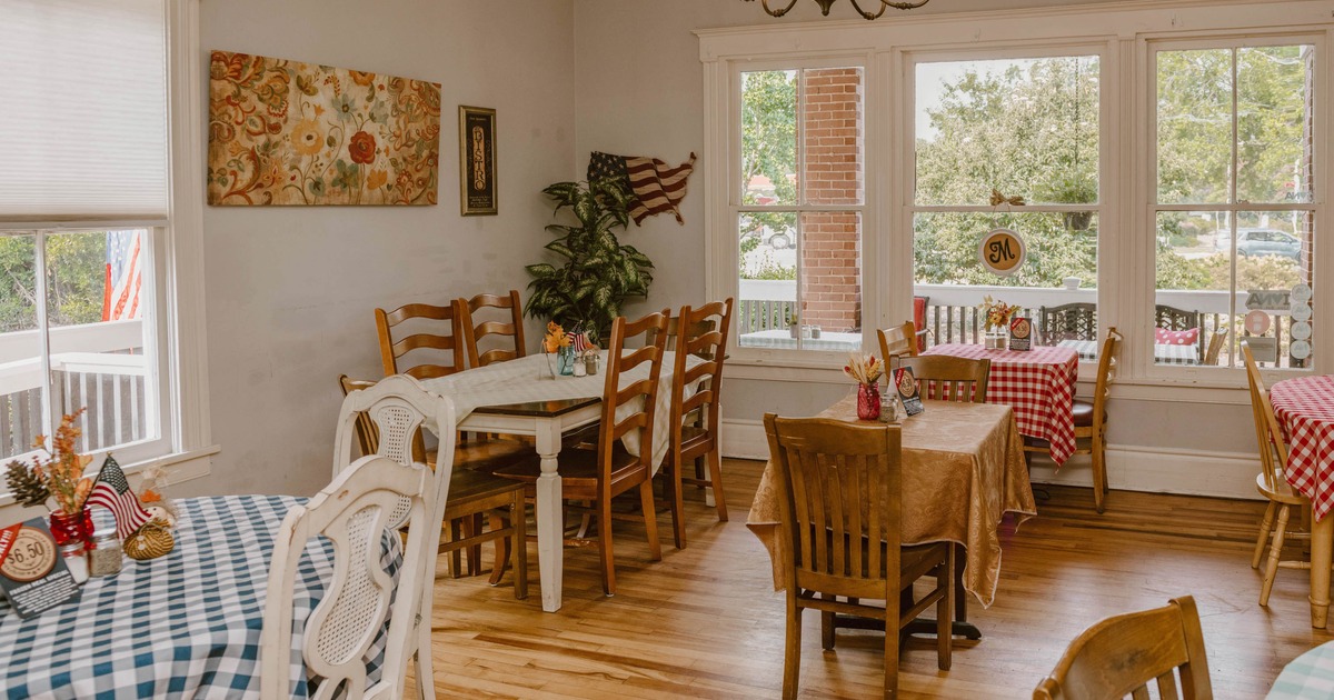 Dining area with patterned tablecloths, wooden chairs, paintings and large windows