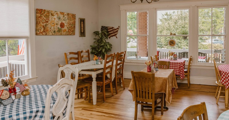 Dining area with patterned tablecloths, wooden chairs, paintings and large windows