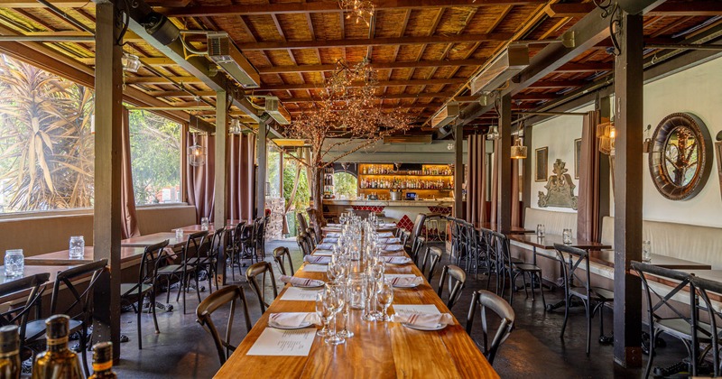 Rustic restaurant interior with a long wooden table, chairs, and a bar in the background