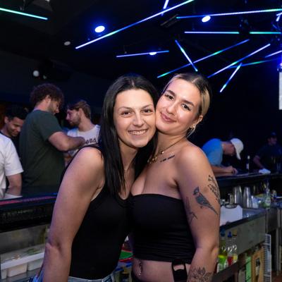 Two staff members pose at a bar with colorful lights overhead and patrons in the background.