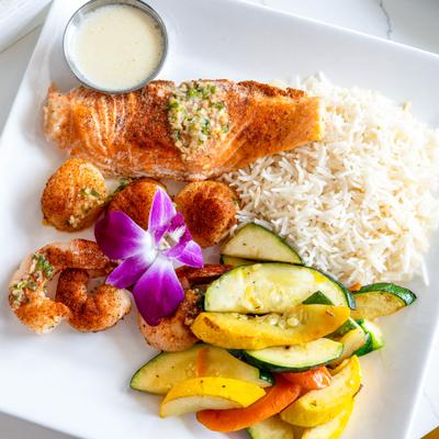 Grilled salmon with shrimp, scallops, rice, sauteed veggies, and sauce on a plate.
