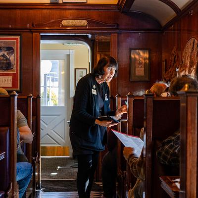Train carriage interior, a staff member takes an order from customers seated in red booths.