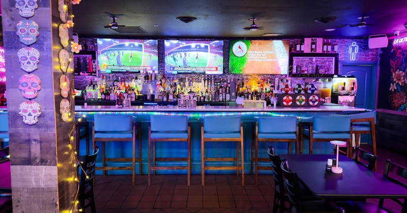 Vibrant bar with colorful lighting, displaying a long counter, bar stools and TVs